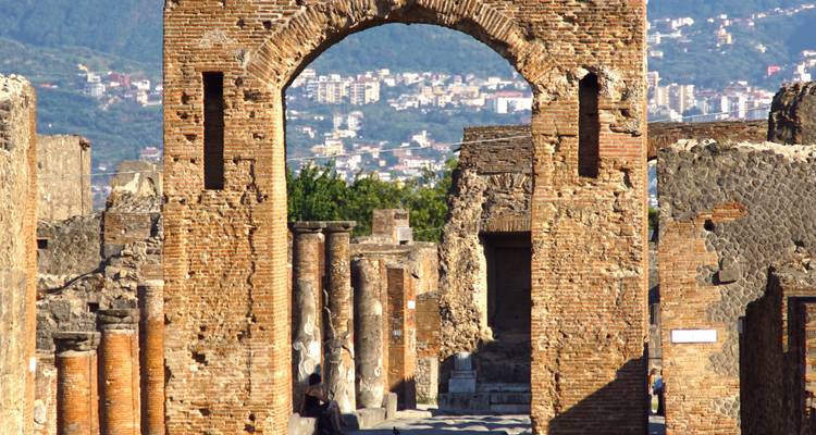 Ancient brick archway and columns in the ruins of Pompeii with a modern city and mountain backdrop under bright blue skies.