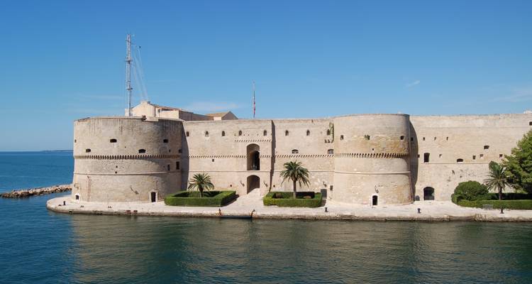 Large stone coastal fortress with palm trees reflected in calm blue water on a clear day.