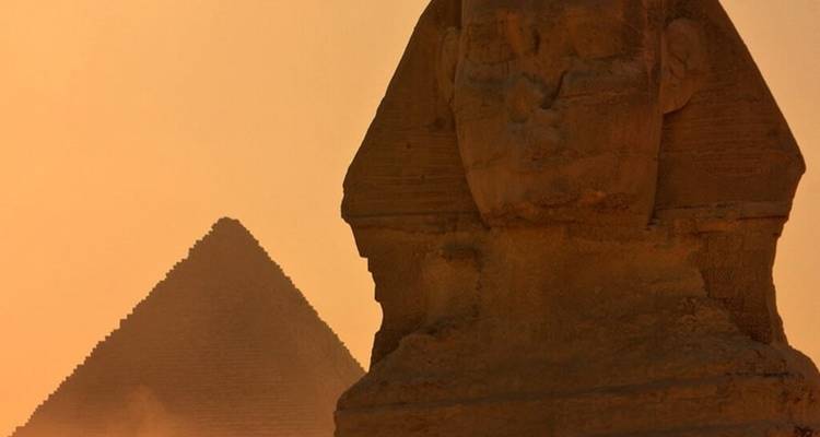 Silhouetted Great Sphinx before hazy pyramid in warm desert sunset light.