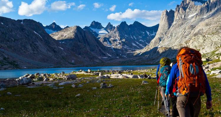 Backpackers trek along a valley floor beside a blue alpine lake and jagged peaks.