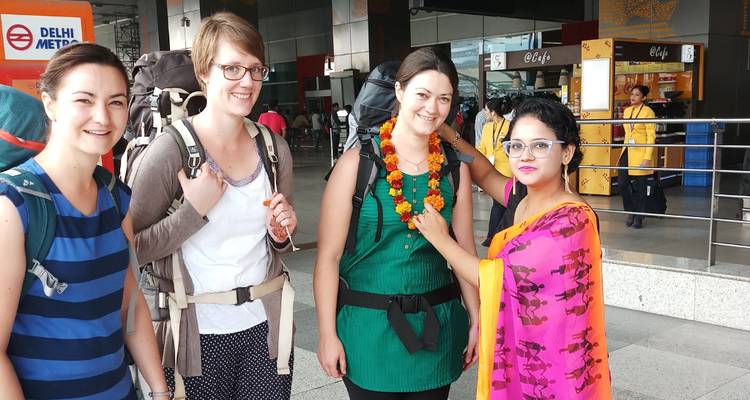 Four travellers at Delhi airport being welcomed with a marigold garland by a hostess in bright saree.