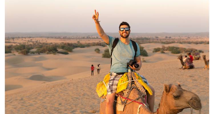 Young man giving a victory sign while riding a camel over golden desert dunes at sunset.