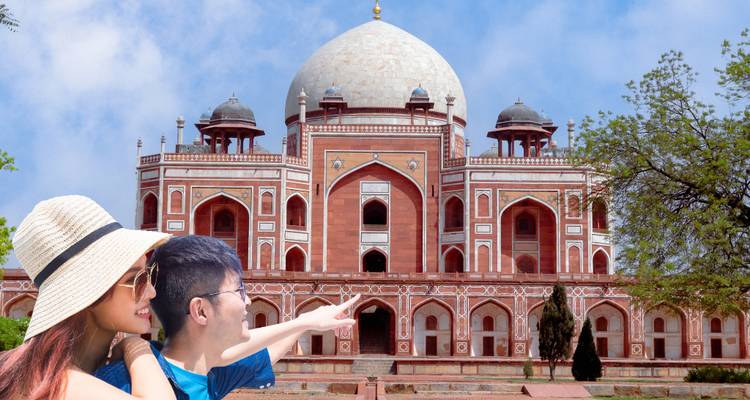 Travelling couple pointing toward the ornate red-sandstone Humayun’s Tomb under a blue sky.