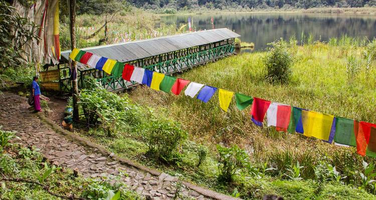 Colorful prayer flags strung beside a wooden bridge over a quiet, reed-lined lake.