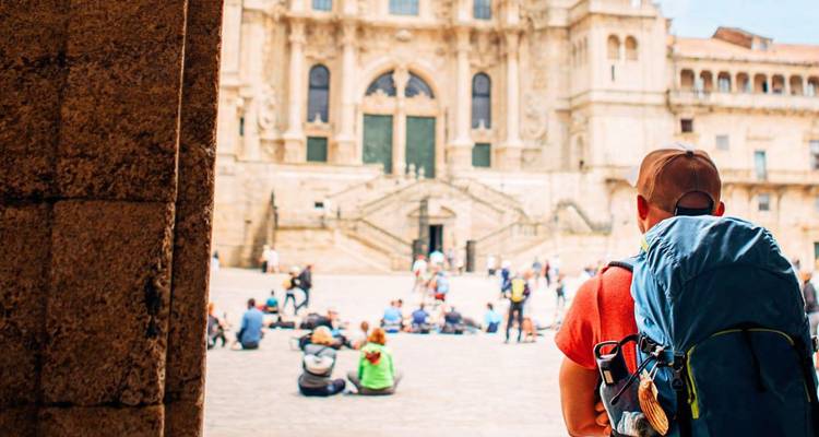 A backpacker with a large pack stands under an archway looking at crowds seated before a grand cathedral plaza.