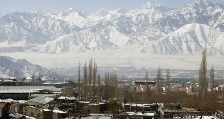 Snow-covered Himalayan mountains rise above a dry valley dotted with village buildings and poplar trees.