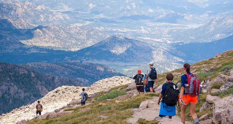 Group of hikers with backpacks descend a rocky mountain trail overlooking expansive valleys.