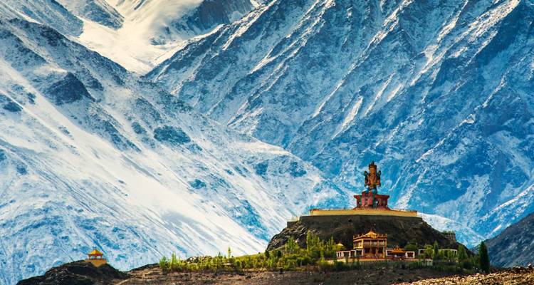 Large golden Buddha statue sits on a pedestal against towering snowy mountain backdrop.
