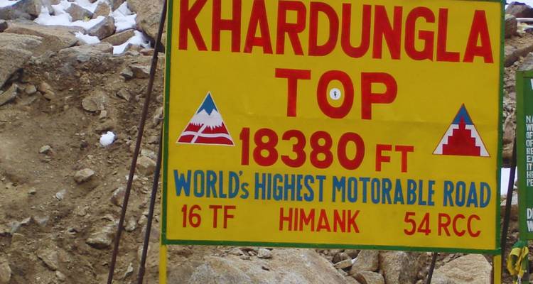 Bright roadside sign marking Khardung La Top surrounded by rocky, snowy mountain terrain.