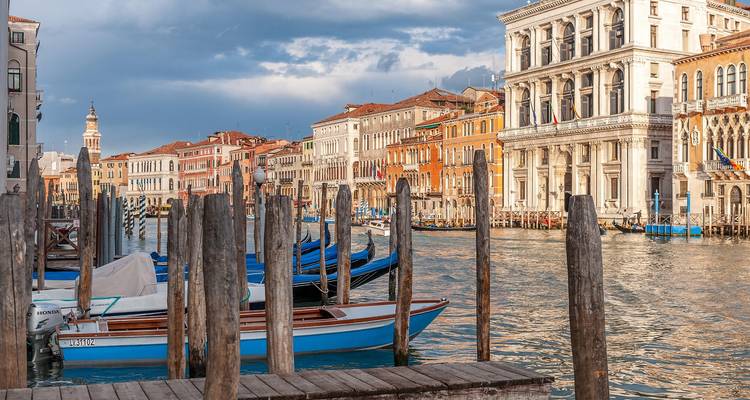 Farbenfrohe Fassaden entlang des Großkanals von Venedig mit vertäuten Gondeln und Holzpfählen unter dramatischem Himmel