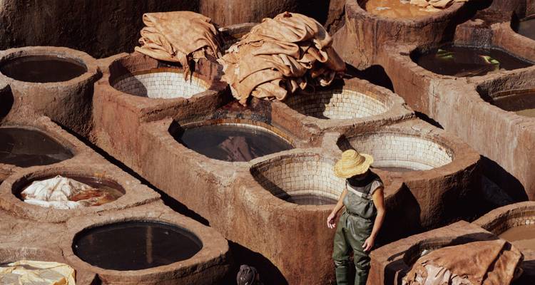 Worker wearing straw hat stands among large circular stone vats of dye at a traditional tannery.