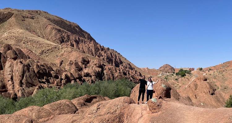 Two hikers pose victoriously atop rugged red rock formations in a desert canyon landscape.