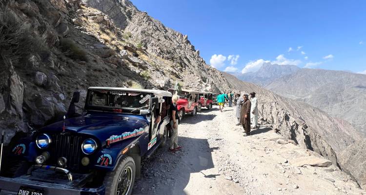 Ligne de jeeps décapotables colorées et de voyageurs se tenant sur une route de montagne étroite et poussiéreuse avec des pics rocheux en arrière-plan.