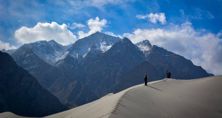 Deux trekkeurs marchant le long d'une crête de dune de sable abrupte avec les pics enneigés du Karakoram en arrière-plan.