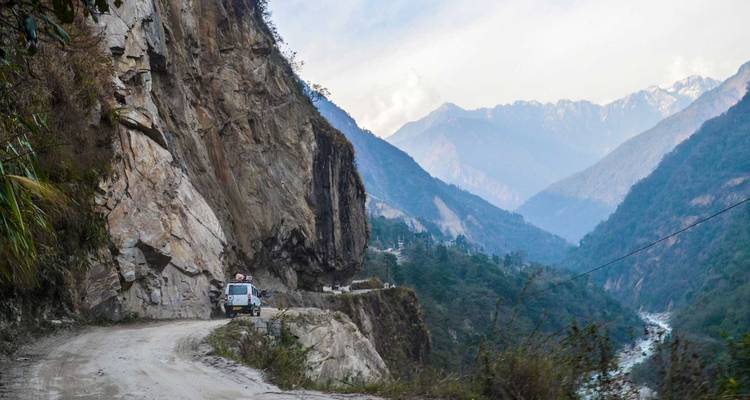 Narrow cliffside road overlooking deep forested valley and distant peaks.