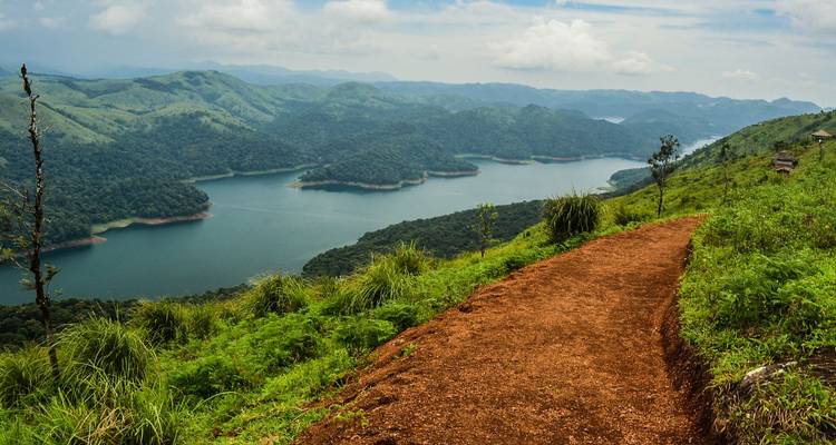 Sentier de randonnée en terre rouge offrant une vue sur un vaste réservoir et des collines boisées.
