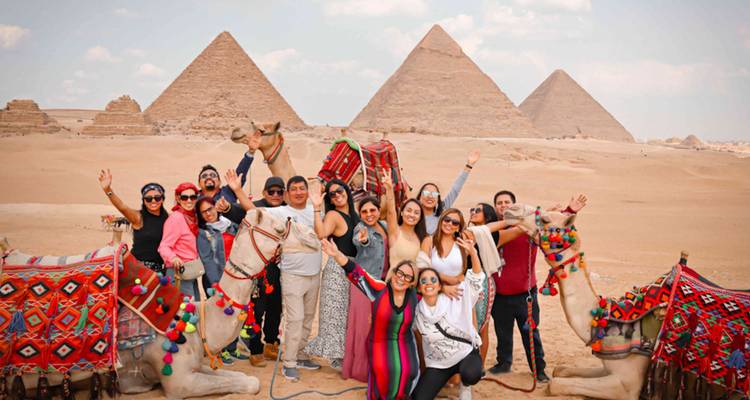 Excited tour group posing with camels in front of the three Great Pyramids in the desert.