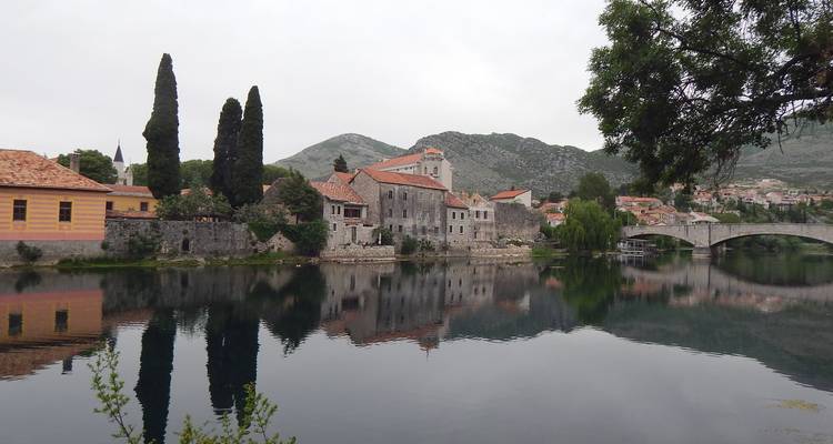 Une ville tranquille au bord d'une rivière avec des maisons en pierre, de grands cyprès et un vieux pont en arche se reflétant dans une eau calme sous un ciel nuageux.