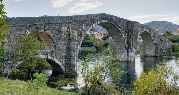 Pont historique en pierre d'Arslanagic avec plusieurs arches traversant une rivière calme, entouré de verdure et de toits de ville éloignés.