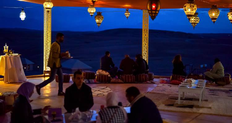 Ambiente de cena nocturna con huéspedes sentados en cojines bajo un dosel iluminado por linternas que da vista a colinas oscuras.