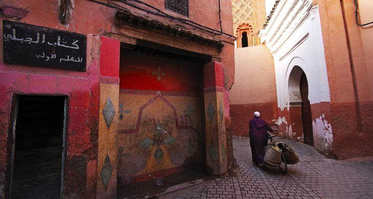 Un hombre con ropa tradicional empuja una bicicleta por un callejón estrecho y colorido con señales en árabe.