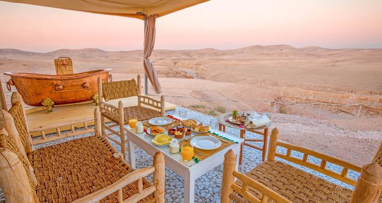 Mesa de desayuno lista con jugos y pasteles bajo una carpa con vistas al desierto ondulante al amanecer.