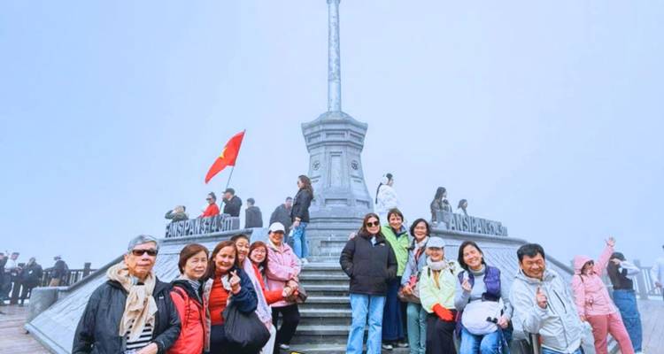 Tour group posing at Fansipan Summit monument on a foggy day holding a flag.