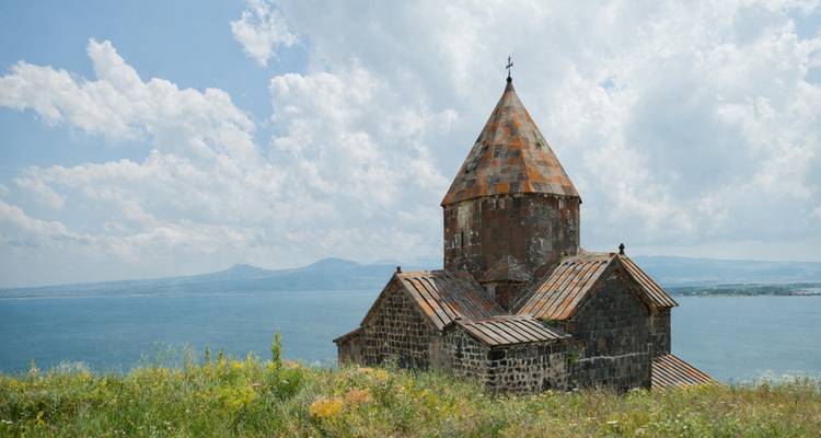 Small stone Sevanavank church perched above the blue waters of Lake Sevan with wildflowers foreground.