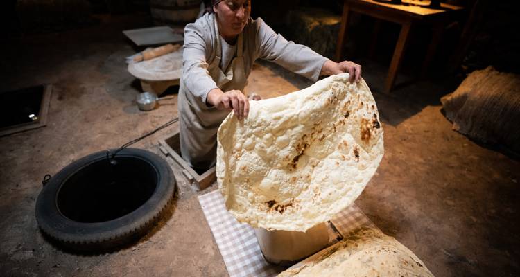 Armenian woman pulls large sheet of traditional lavash bread from a clay oven in rustic room.