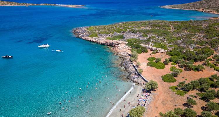 Vue aérienne de la baie turquoise avec des baigneurs, une plage de sable et une presqu'île rocheuse verdoyante.