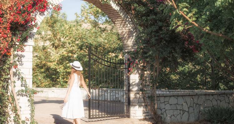 Femme vêtue d'une robe blanche et d'un chapeau se promenant sous une arche de jardin parsemée de lumière solaire et ornée de vignes en fleur.