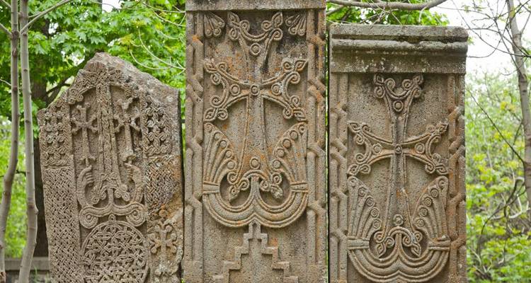 Three intricately carved Armenian khachkar stone crosses standing among green trees.