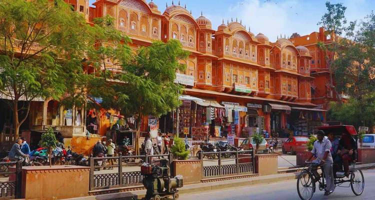 Bustling Jaipur street lined with ornate pink facades, vendors and rickshaws under blue skies.