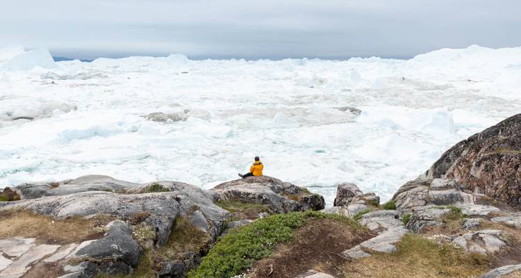 Ein einzelner Reisender in gelbem Parka meditiert neben der riesigen Eisfläche des Ilulissat-Eisfjords.