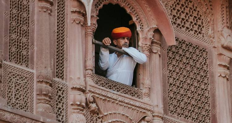 Ein Mann in orangefarbenem Turban blickt aus einem aufwendig geschnitzten Steinenfenster eines historischen Palastes.