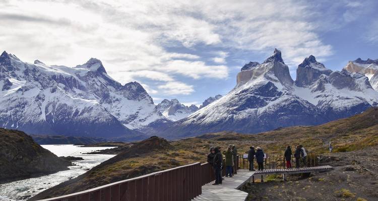 Small group on a wooden boardwalk taking in sweeping snowy mountain vistas and rushing river below.