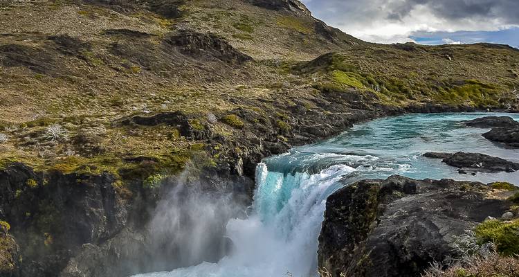 Powerful turquoise waterfall plunges into a mist-filled gorge amid rugged Patagonian terrain.