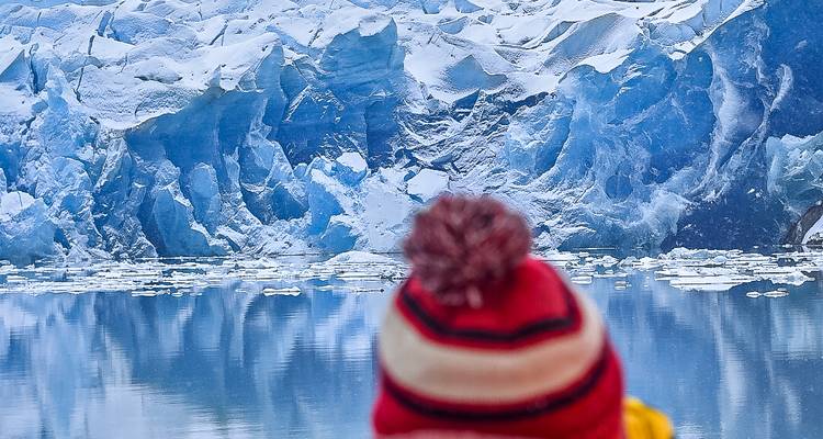 Traveler in red wool hat gazes at towering blue glacier walls reflected in still water.