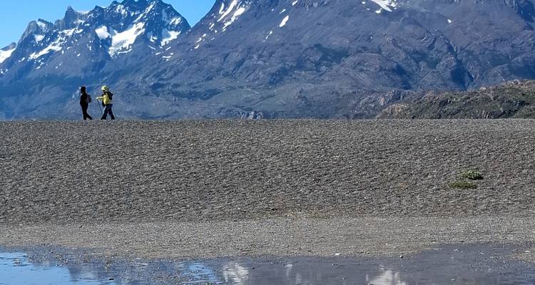 Two hikers traverse a vast gravel plain with towering snow-peaked mountains behind.