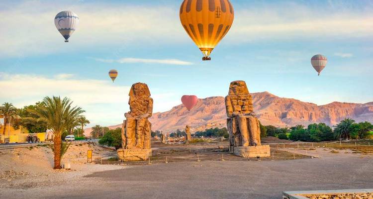 Hot air balloons floating above the Colossi of Memnon with desert mountains behind.