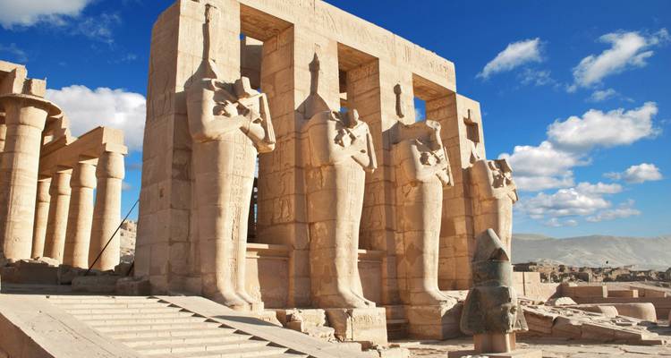 Row of massive standing statues inside an open-air Egyptian temple beneath bright sky.