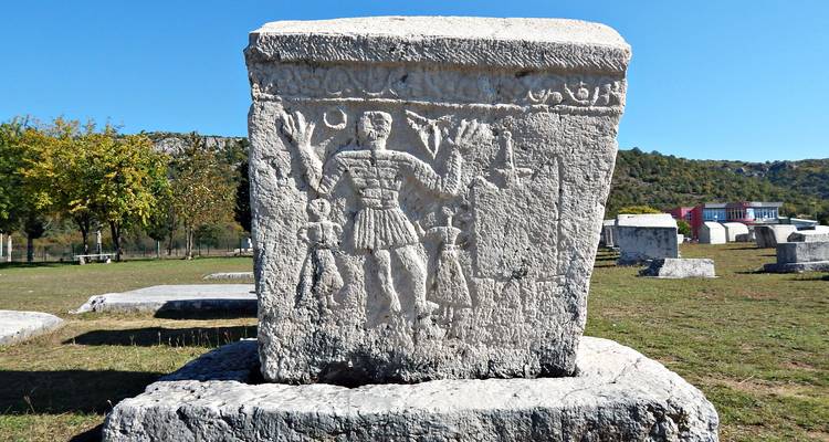 Una antigua lápida de piedra caliza tallada stećak se encuentra en un campo arqueológico abierto bajo cielos despejados.