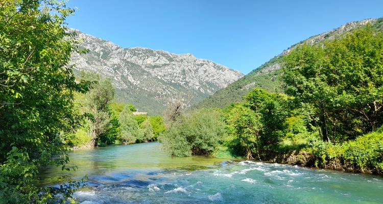 Río de aguas turquesas cristalinas que serpentea a través de un frondoso bosque en la base de montañas rocosas en un día soleado.