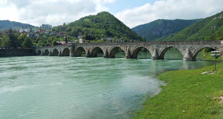 Icónico puente de piedra otomano con múltiples arcos que atraviesa un amplio río verde rodeado de colinas boscosas.