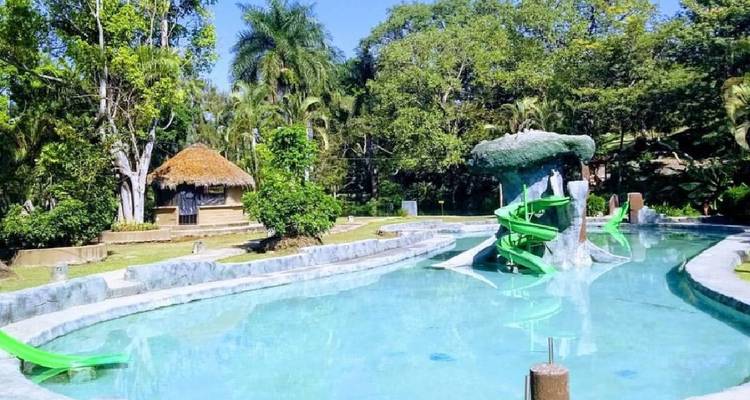 Außenpool-Komplex mit leuchtend grünen Wasserrutschen und Palapa-Hütte, umgeben von tropischen Bäumen.