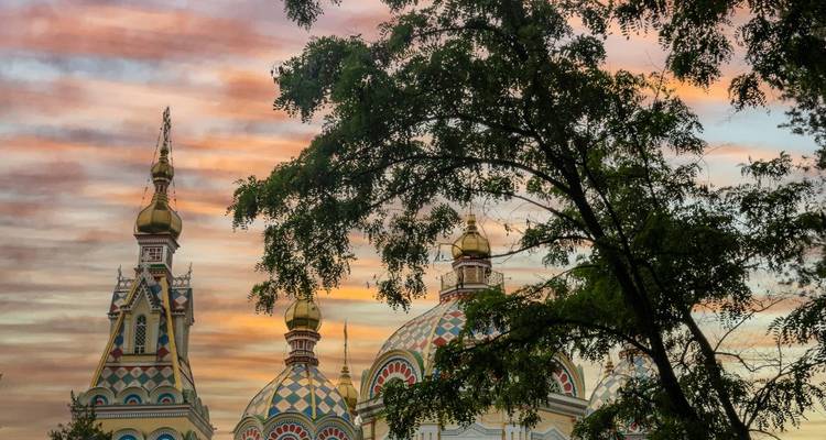 Golden evening sky with pastel clouds framing the ornate domes of Zenkov Cathedral partly hidden behind leafy tree branches.