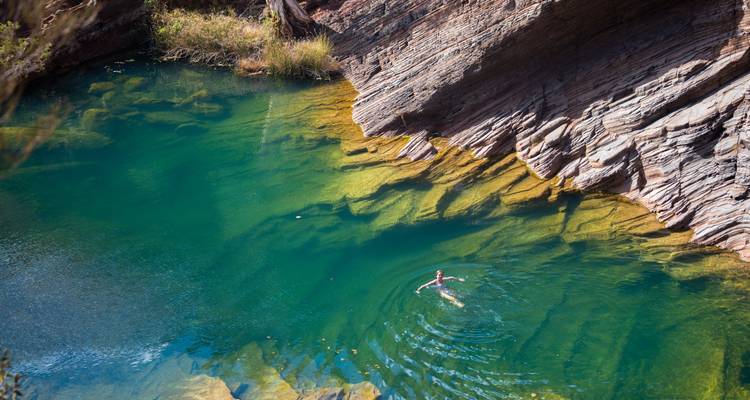 A swimmer floats in an emerald green natural pool bordered by striking banded rock walls.
