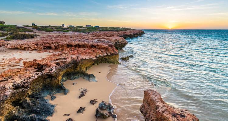 Turquoise waves lap against rugged red-rock cliffs on Western Australia’s Ningaloo Coast at sunset.