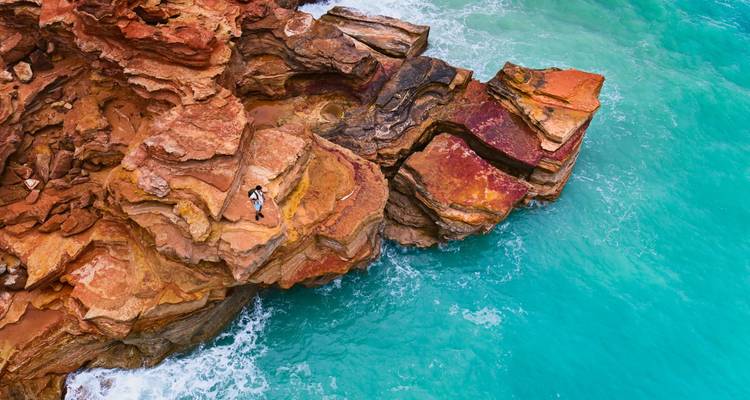 Aerial view of rugged red sandstone cliffs meeting bright turquoise sea, with a lone sunbather on the rocks.