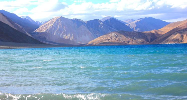 Brilliant blue waters of Pangong Tso stretch toward multicolored barren mountains under a bright sky.